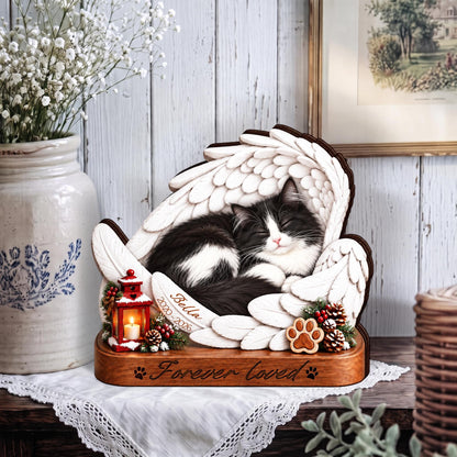 A memorial plaque featuring a black and white cat sleeping within white angel wings. The base is wood-toned with the text 'Forever loved' and paw prints. A small red lantern, pinecones, and a name tag reading 'Bella 2020-2026' are included.