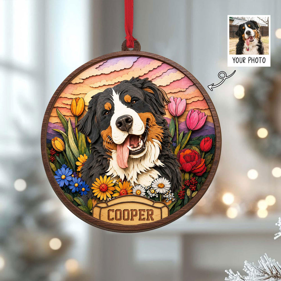 A round wooden ornament featuring a layered illustration of a Bernese Mountain Dog with its tongue out, surrounded by colorful tulips and daisies against a sunset sky. A banner at the bottom displays the name COOPER in capital letters.