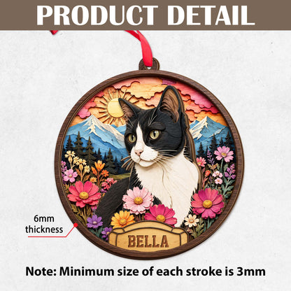 A circular wooden ornament featuring a layered 3D design of a black and white cat surrounded by colorful flowers, mountains, and a sun. A nameplate at the bottom reads BELLA. The ornament has a 6mm thickness and a red hanging ribbon.