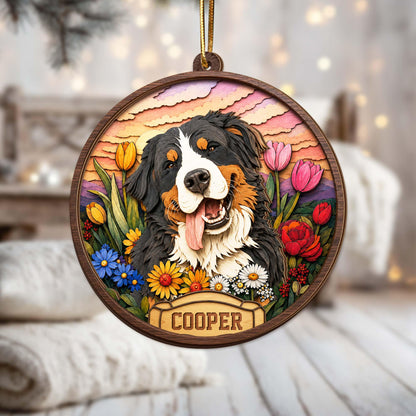 A round wooden ornament featuring a layered, multi-dimensional illustration of a Bernese Mountain Dog surrounded by colorful tulips, daisies, and roses against a sunset sky. A banner at the bottom displays the name COOPER.