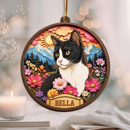 A circular wooden ornament featuring a layered 3D-style illustration of a black and white cat. The background displays a sun, blue mountains, and a forest. The foreground includes colorful flowers and a nameplate with the text BELLA.