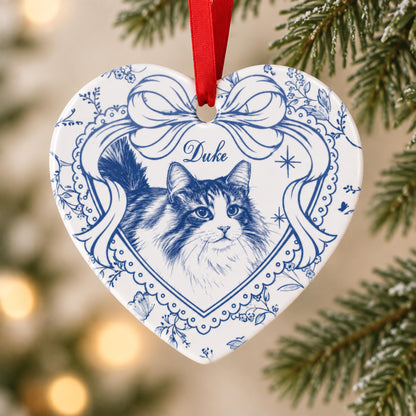 A heart-shaped white ceramic ornament featuring a blue ink-style illustration of a long-haired cat. The design includes a decorative blue ribbon border, floral accents, and the name Duke printed in blue script, suspended by a red ribbon.
