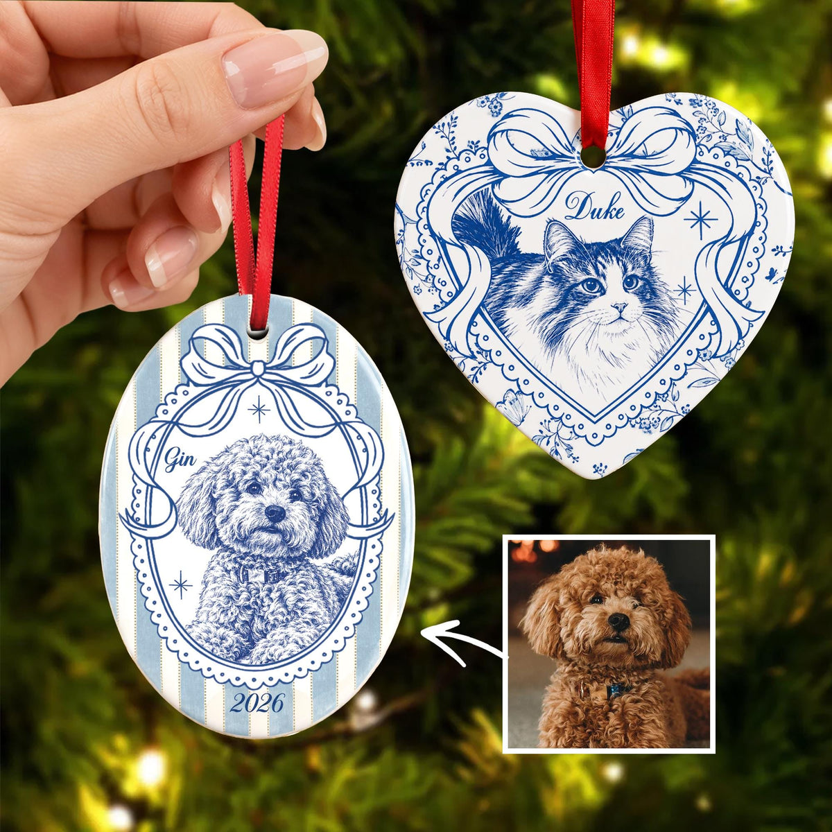 Two ceramic ornaments with blue and white illustrations. One oval ornament features a curly-haired dog labeled Gin and the year 2026. The heart-shaped ornament features a long-haired cat labeled Duke. Both hang from red ribbons.