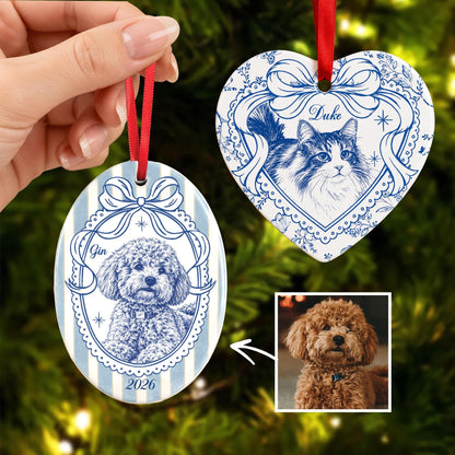 Two ceramic ornaments with blue and white illustrations. One oval ornament features a curly-haired dog labeled Gin and the year 2026. The heart-shaped ornament features a long-haired cat labeled Duke. Both hang from red ribbons.