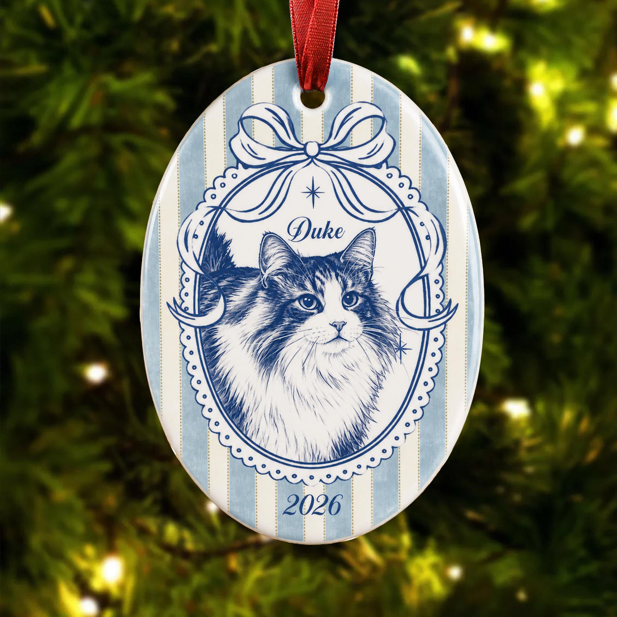 An oval ceramic ornament featuring a blue and white striped background. A central oval frame with a bow contains a detailed blue line drawing of a long-haired cat. The text "Duke" appears above the cat and "2026" below it.