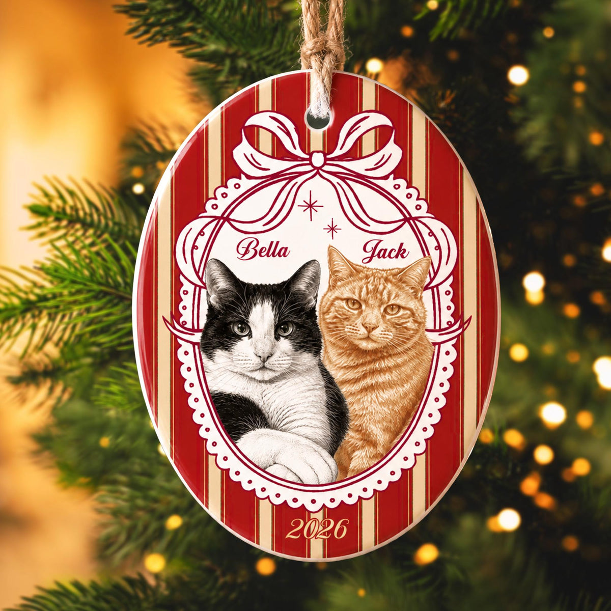 An oval ceramic Christmas ornament featuring a red and cream striped background. The center displays a white scalloped frame with a bow, containing illustrations of a black and white cat named Bella and an orange tabby cat named Jack, with the year 2026 printed at the bottom.
