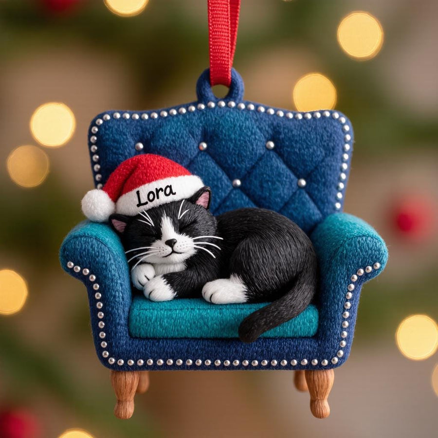 A Christmas ornament featuring a black and white cat wearing a Santa hat that says "Lora" resting on a blue armchair. The ornament has a red ribbon for hanging and is set against a blurred background of Christmas lights.