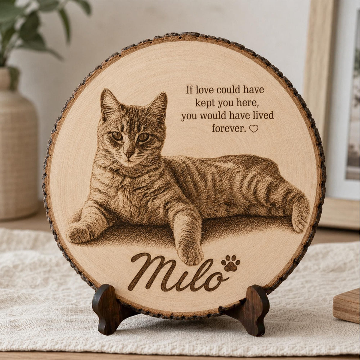 A personalized round natural wood slice plaque featuring a detailed engraved-style portrait of a tabby cat named Milo. The design includes the memorial quote “If love could have kept you here, you would have lived forever,” a small heart and paw print, and Milo’s name in elegant script. The rustic bark-edged wood slice is displayed on a dark wooden stand in a warm neutral home setting.