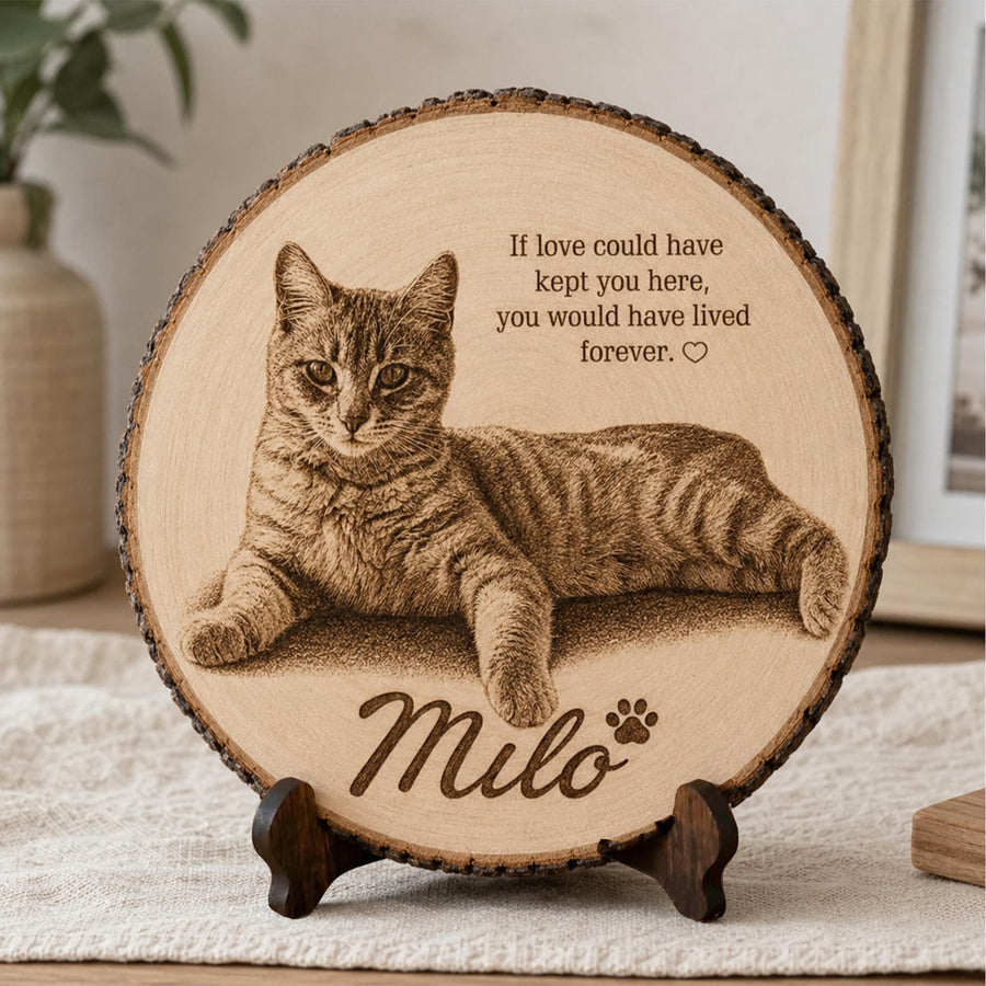 A personalized round natural wood slice plaque featuring a detailed engraved-style portrait of a tabby cat named Milo. The design includes the memorial quote “If love could have kept you here, you would have lived forever,” a small heart and paw print, and Milo’s name in elegant script. The rustic bark-edged wood slice is displayed on a dark wooden stand in a warm neutral home setting.