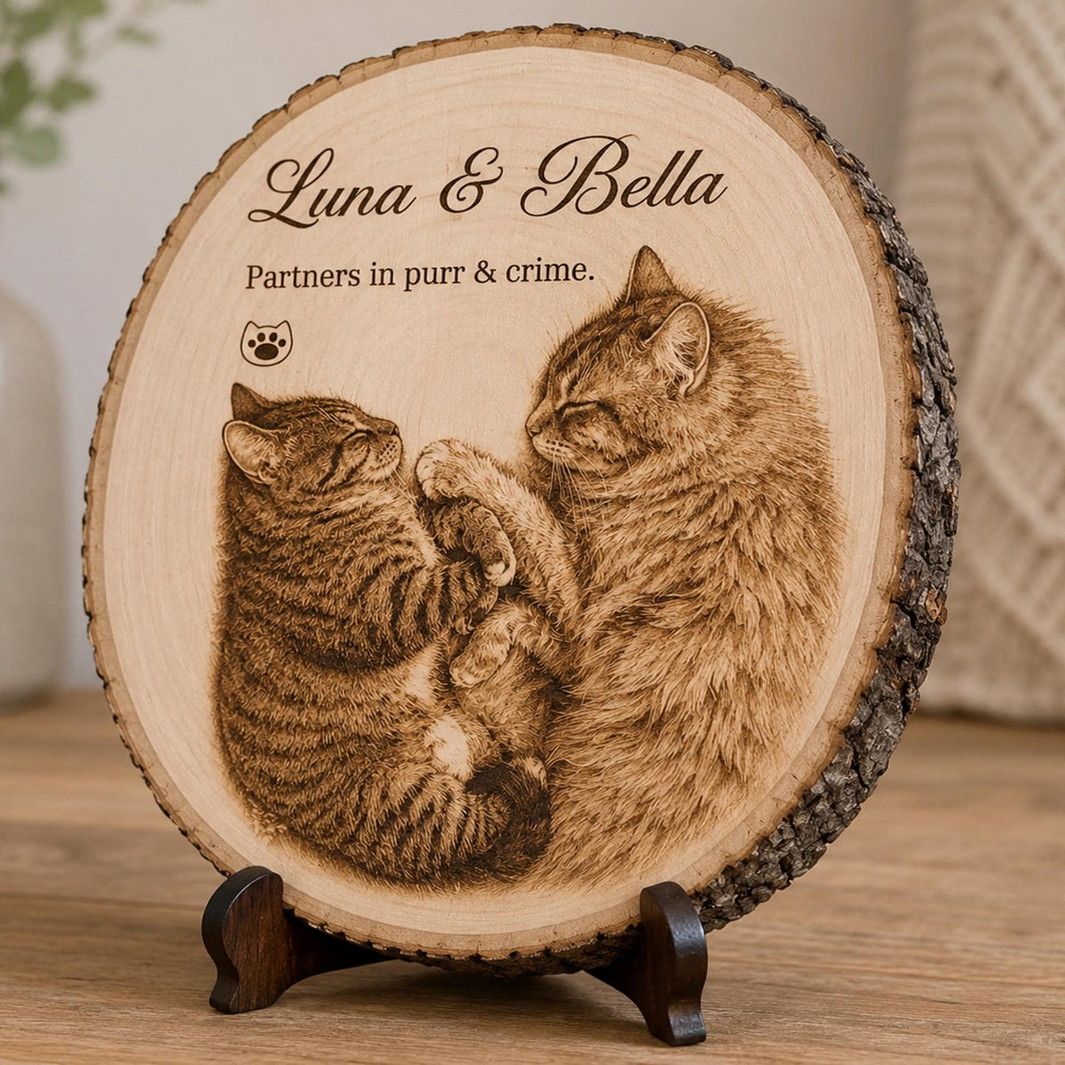 A circular wooden plaque with natural bark edges, featuring a laser-engraved image of two cats cuddling. The text reads "Luna & Bella" and "Partners in purr & crime." with a small paw print icon. It rests on a dark wooden display stand.