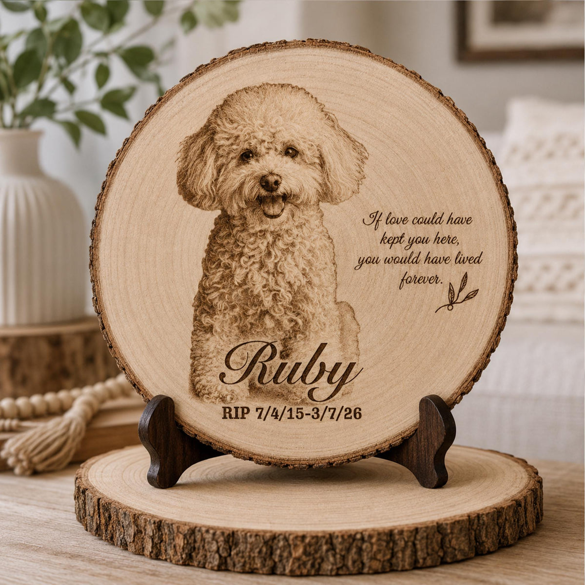 A circular wood slice memorial plaque featuring a laser-engraved portrait of a curly-haired dog. The text reads: If love could have kept you here, you would have lived forever. Ruby, RIP 7/4/15-3/7/26. The plaque rests on a dark wooden stand.