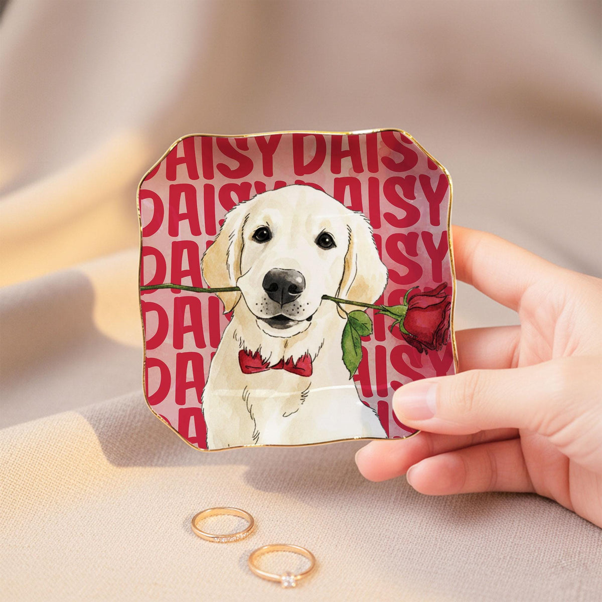 A square ceramic trinket dish with wavy edges and a gold-painted rim. The surface features a repeating red text pattern reading DAISY, a central illustration of a cream-colored dog wearing a red bow tie, and a red rose held in its mouth.