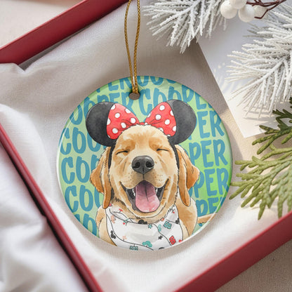 A round ceramic ornament featuring a watercolor illustration of a golden retriever wearing black mouse ears with a red and white polka dot bow and a white bandana with small colorful icons, set against a light green background.