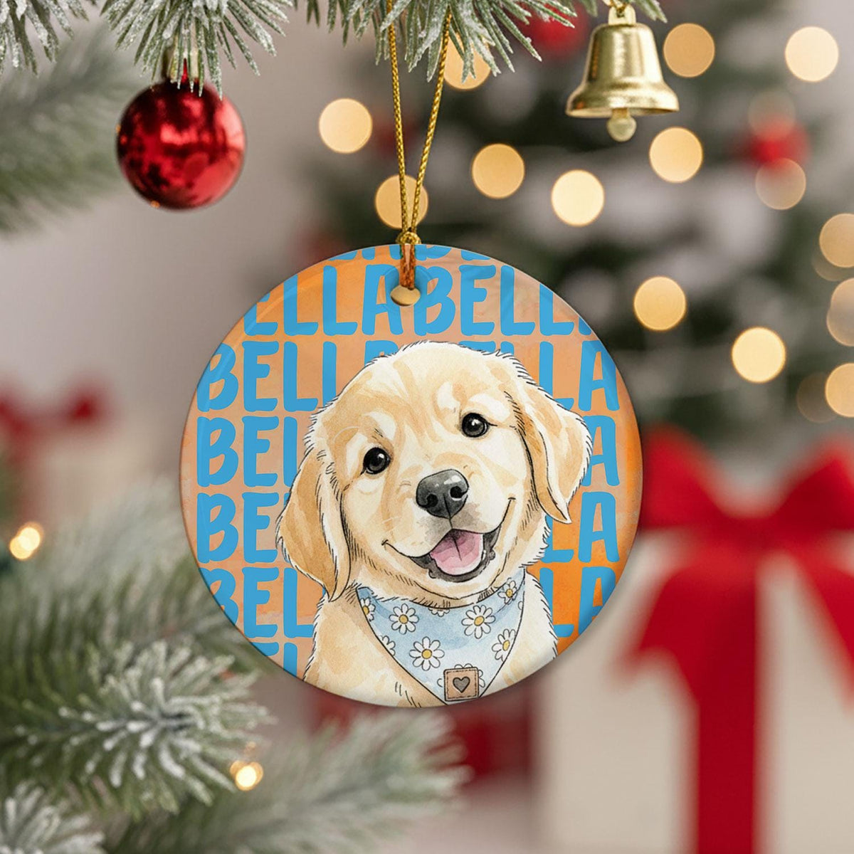 A round ceramic ornament featuring a golden retriever puppy illustration wearing a light blue bandana with white daisy flowers. The background displays the name Bella repeated in blue text over an orange backdrop, suspended by a gold cord.