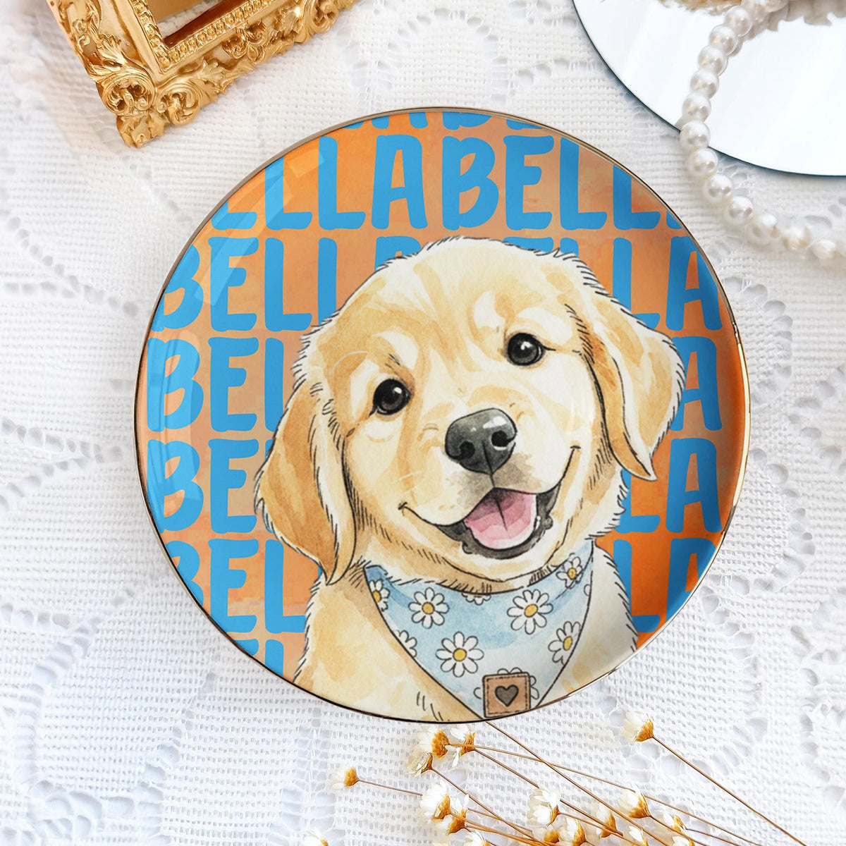 A round ceramic plate featuring a golden retriever puppy illustration wearing a light blue bandana with a white daisy pattern. The background consists of a repeating orange and blue text pattern that reads BELLA repeatedly.