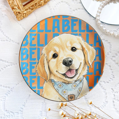 A round ceramic plate featuring a golden retriever puppy illustration wearing a light blue bandana with a white daisy pattern. The background consists of a repeating orange and blue text pattern that reads BELLA repeatedly.